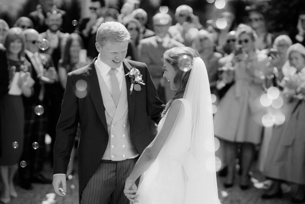 Timeless black and white moment of a bride and groom smiling at each other, surrounded by guests and floating bubbles after the ceremony.
